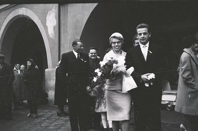 Black-and-white funeral procession outside a building with arched entryways. Central figures: woman in formal dress holding f...