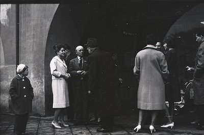 Mid-20th century street scene under arched stone passageway. Group of adults in formal attire—men in suits, women in knee-len...