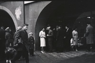 Black-and-white street scene under arched concrete overpass, likely 1950s–1960s. Group of adults and children in mid-century ...
