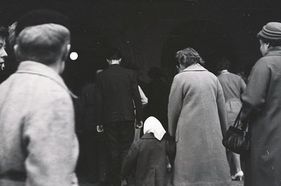 A candid black-and-white street scene from the 1950s–60s, featuring a diverse group of people in mid-20th-century attire. A w...