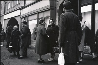 Black-and-white street scene featuring mid-20th century European urban life. Crowd of individuals in heavy winter coats, carr...