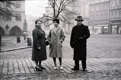 Three individuals pose on cobblestone street in 1950s European city square. Women wear knee-length coats with structured shou...