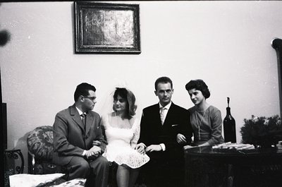 Mid-20th century wedding portrait in a formal indoor setting. Four individuals pose: bride in lace dress with veil, groom in ...