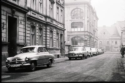 Classic 1950s European street scene with vintage cars parked along a cobblestone road. Architectural details include ornate f...