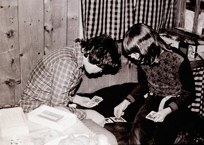 Two children playing cards on a couch in a modest, wood-paneled interior, likely mid-20th century. The woman’s checkered blou...