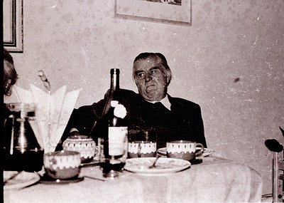Mid-20th century indoor dining scene featuring an elderly man in formal attire holding a wine bottle. Table set with vintage ...