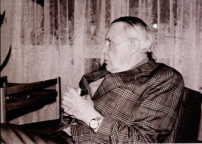 Mid-20th century portrait of an elderly man in a plaid sweater, seated indoors with a wooden chair. Faded wallpaper and vinta...