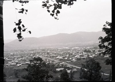 Aerial black-and-white view of a densely packed village nestled between rolling hills, framed by tree branches. Low-rise buil...