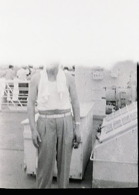 Mid-20th century man in loose trousers and tank top stands on a dock, holding a small object. Industrial port setting with vi...