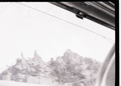 Vintage black-and-white shot of rugged alpine peaks viewed through a car window, suggesting mid-20th century travel. Distorte...