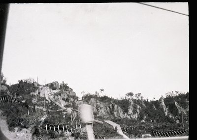 Black-and-white rural landscape featuring a dirt road winding through rocky terrain with sparse vegetation. A cylindrical wat...