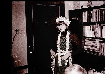 Vintage black-and-white photo of a young girl in a maid’s uniform—lace apron, blouse, and cap—posing indoors beside a bookshe...