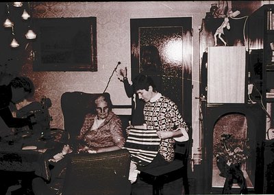 Vintage sepia-toned indoor portrait of two women in a modest living space, likely 1960s–1970s. One woman, seated on a woven c...