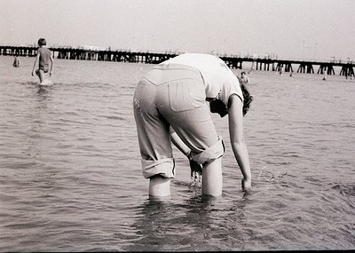 Black-and-white beach scene from mid-20th century (1950s–1960s), showing women in knee-length swimsuits wading in shallow wat...