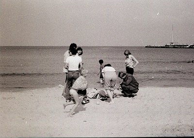 Group of people on a sandy beach, mid-20th century. One person lies on sand while others kneel or stand around, likely during...