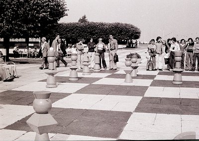 Large-scale outdoor chess game on a checkerboard-patterned plaza with ceramic chess pieces. Mid-20th century attire suggests ...