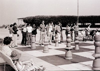 Large outdoor chess set with 1970s-style attire; people engaged in casual play. Urban park setting with trees and paved area.