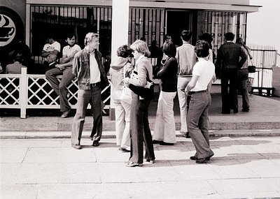 Group of young adults in 1970s streetwear—bell-bottoms, wide lapels, and casual poses—gathering outside a building with wroug...