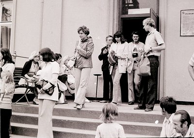 1970s street scene outside a Soviet-era building, featuring a diverse group of people in casual attire—men in button-downs, w...
