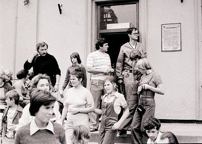 A candid black-and-white street scene from the **1970s**, featuring a diverse group of young adults and children gathered out...