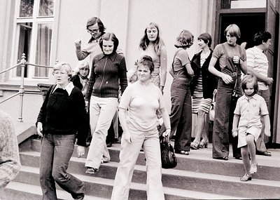 Group of 12 people descending grand stone staircase in 1970s European urban setting. Women in wide-leg trousers, men in short...