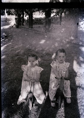 Vintage black-and-white photo of two young children in matching overalls, riding a tandem tricycle on a paved driveway. Trees...