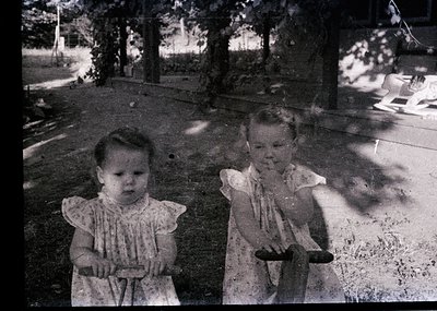 Vintage black-and-white photo of two young girls in early 20th-century dresses, posing outdoors on a paved path. Trees and gr...