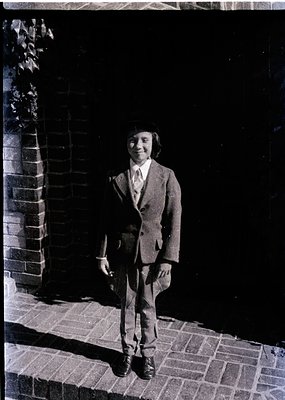 Young man in a 1960s-70s suit poses outdoors near brick wall and potted plant. Classic tailored jacket, dress shirt, and trou...