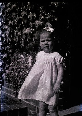 Vintage black-and-white portrait of a young girl in a 1950s-style dress with a bow, standing on stone steps beside leafy foli...