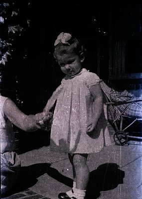 Vintage black-and-white photo of a young child in a knee-length dress with short sleeves, standing outdoors near a pram. The ...