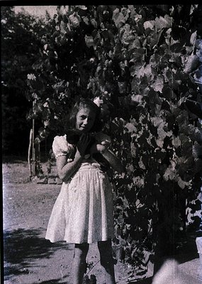 Young girl in vintage dress hugs a plush toy, standing beside leafy vine-covered wall. Mid-20th century rural or suburban set...