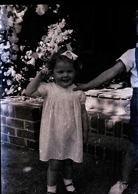 Vintage sepia-toned photo of a young girl in a short-sleeved dress with a bow, standing beside a brick oven or stove. Her joy...