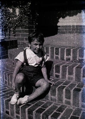 Vintage black-and-white photo of a boy in overalls and white shoes sitting on brick steps, smiling directly at the camera. Mi...
