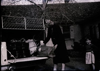 Mid-20th century black-and-white photo: Woman in dark coat and headscarf unloading a large basket from a vintage truck, while...