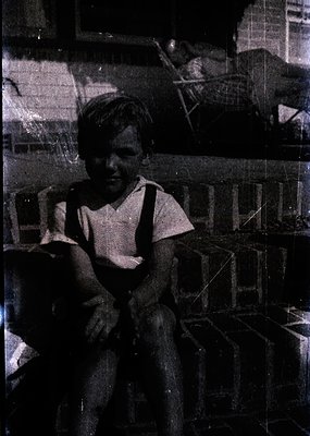 Vintage black-and-white photo of a young boy in suspenders, seated on a wooden chair indoors. Stacked crates and shelves with...
