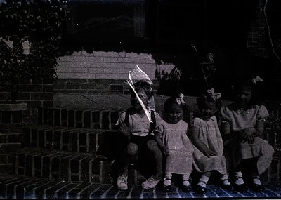 Four children pose on brick steps, holding kites in mid-19th to early 20th century attire—long dresses, bows, and cardigans. ...
