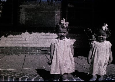 Two young girls in early 20th-century dresses with bows pose outdoors on concrete steps. Sepia-toned vintage photo suggests e...