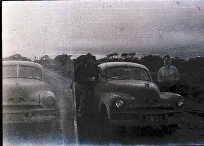 Vintage black-and-white photo of two classic cars parked on a rural road, likely 1950s. A man leans against the rear of a sed...