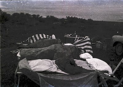 Vintage black-and-white photo of a makeshift outdoor kitchen setup with large metal pots, striped cloths, and a vintage truck...