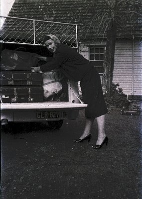 Mid-20th century woman in 1950s-style dress and high heels unloads luggage from a vintage truck with German license plate. Re...