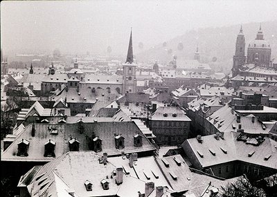 Aerial view of a historic European cityscape blanketed in snow, showcasing dense, low-rise architecture with steep slate roof...