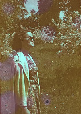 Vintage sepia-toned portrait of a woman in a floral dress, standing outdoors with blurred greenery background. Likely 1950s–1...