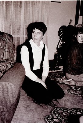 Vintage black-and-white interior shot of a woman in a cropped sweater vest and high-waisted trousers, seated on a patterned r...