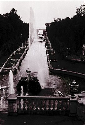 Grand Baroque-style fountain with cascading tiers and central jet, flanked by symmetrical basins. Lush greenery and stone bal...