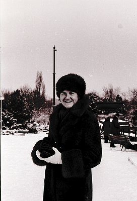 Black-and-white winter portrait of a woman in a fur-lined coat and hat, holding a fur-trimmed glove. Snow-covered park bench ...