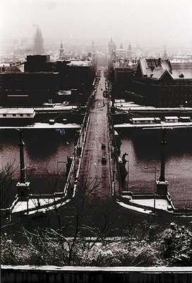 Aerial view of a snow-covered urban canal in winter, likely Budapest. The frozen Danube River splits the cityscape, flanked b...