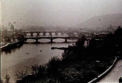 Vintage black-and-white view of a multi-arched stone bridge spanning a river, flanked by sparse winter trees and urban buildi...