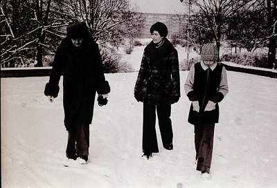 Three individuals in winter attire walk through snow-covered ground, likely a park or residential area. The woman on the left...