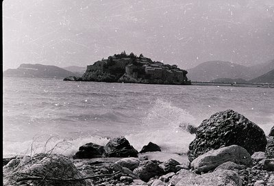 Black-and-white coastal scene featuring a fortified island structure atop rugged cliffs, surrounded by choppy waters and rock...