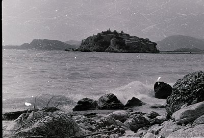 Historic coastal fortress perched on rocky islet, surrounded by turbulent waves. Mid-20th century black-and-white photograph ...